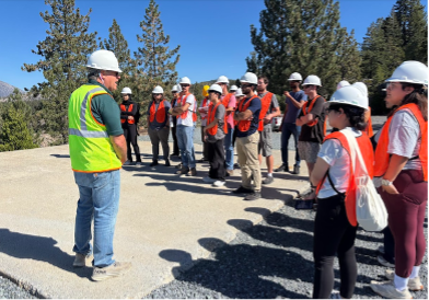 Professor in a green vest meets with students in orange vests.